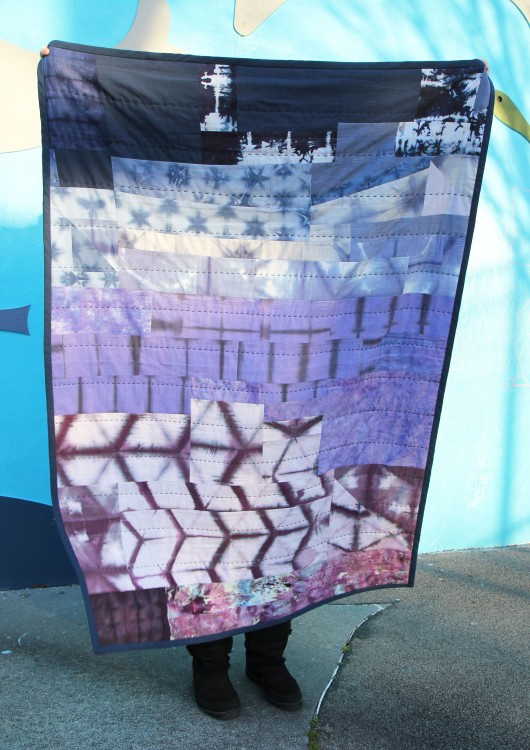 person holding up colorful ombre patchwork quilt outside in front of a wall with a mural on it